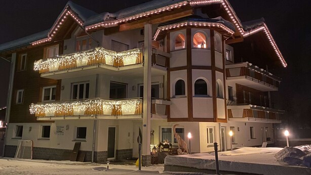 A multi-story hotel with balconies adorned with string lights, a snow-covered ground, and a central tower with arched windows. - Mountain Stay With Balcony (Saas Grund)
