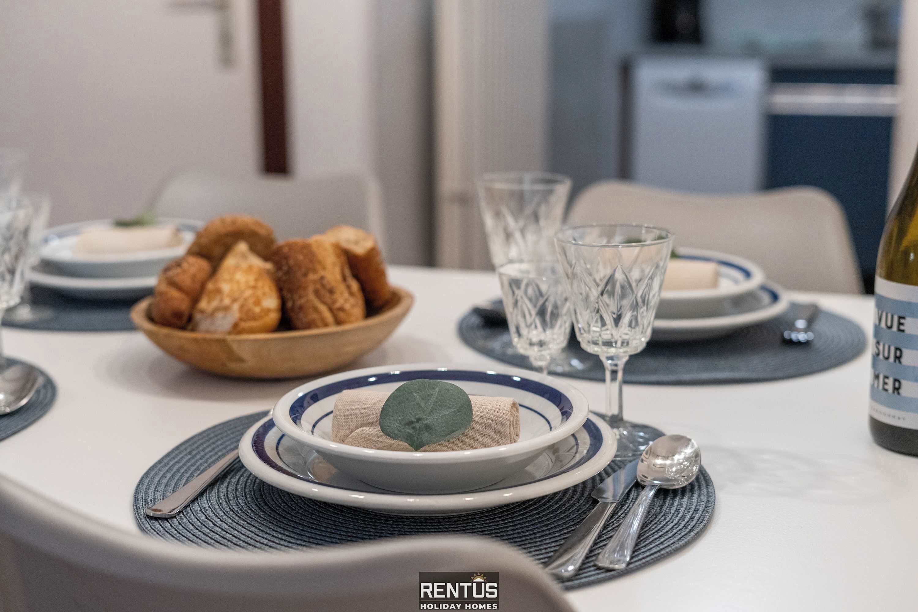 Una tavola imbandita con piatti, bicchieri e una bottiglia, oltre a una ciotola di pane e un tovagliolo su un sottopiattino.