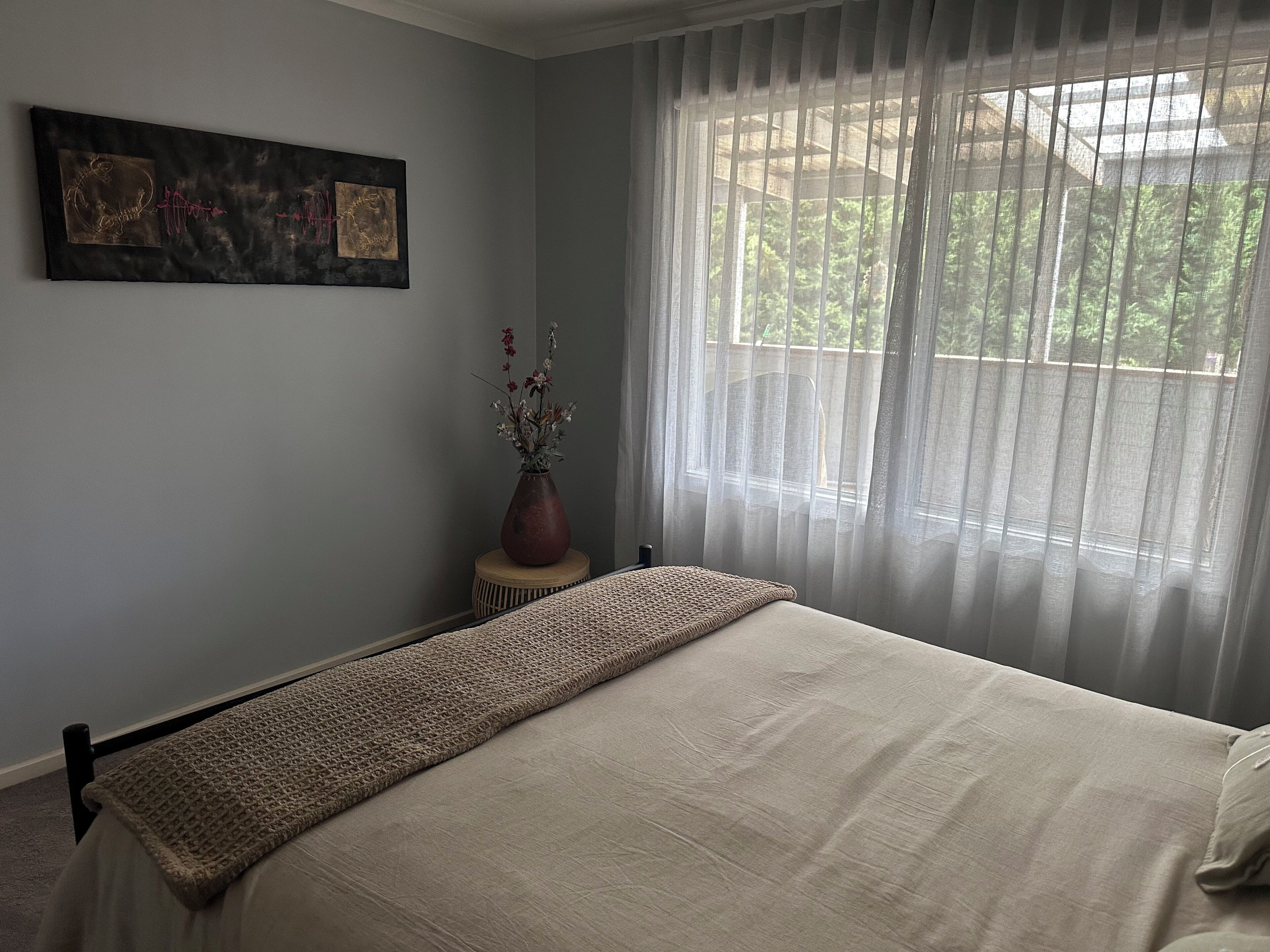 A bedroom with a large bed, a window with sheer curtains, a small table with a vase of flowers, and a wall-mounted artwork.