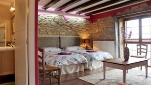 Un dormitori amb paret de pedra, dos llits, una cadira de fusta, una taula i una finestra amb vistes.