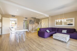 Un salon moderne comprenant un coin repas, un canapé violet, un pouf blanc et un escalier.