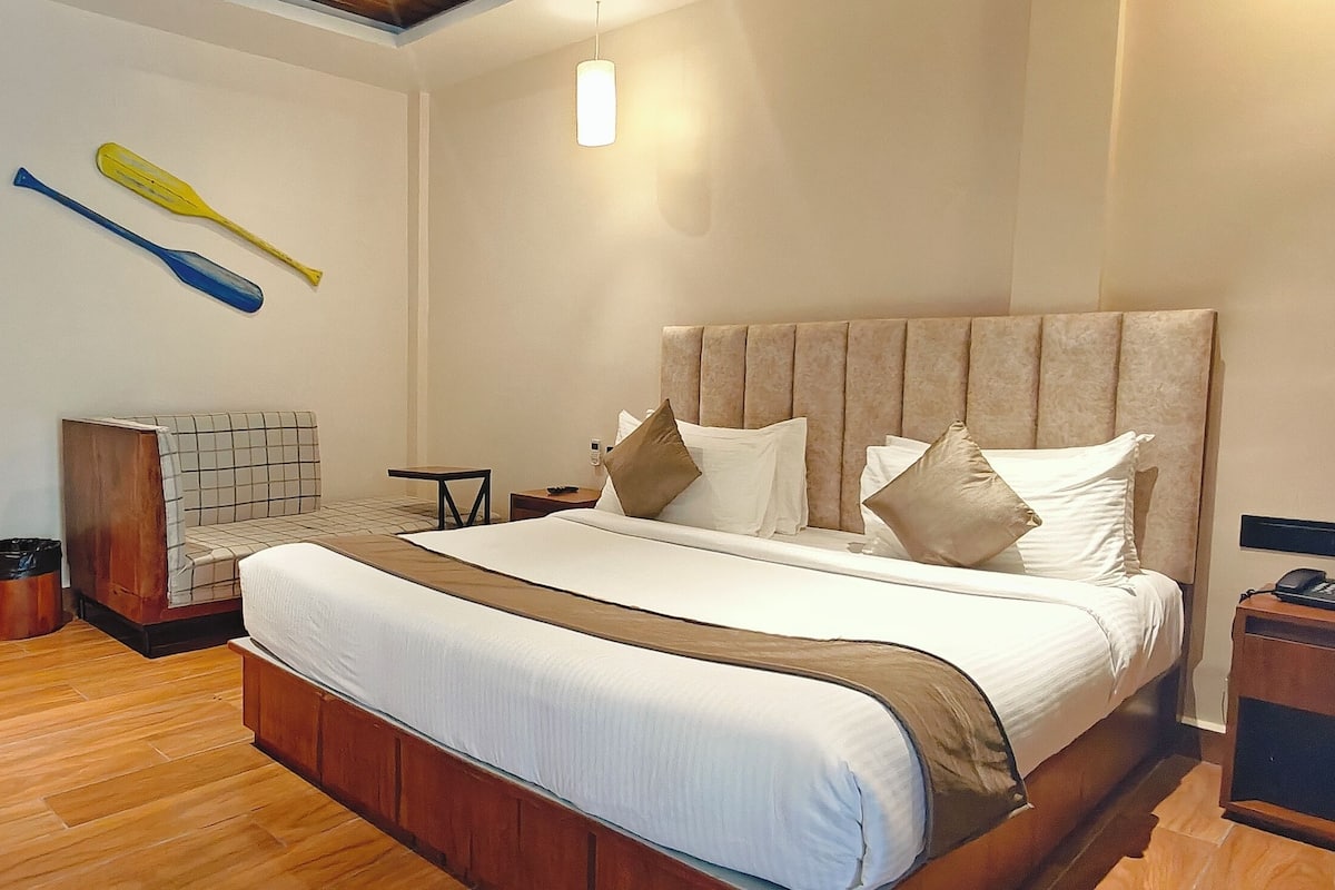 A hotel room with a large bed, a bedside table, a nightstand, a wall-mounted TV, and a small seating area with a bench and a table.
