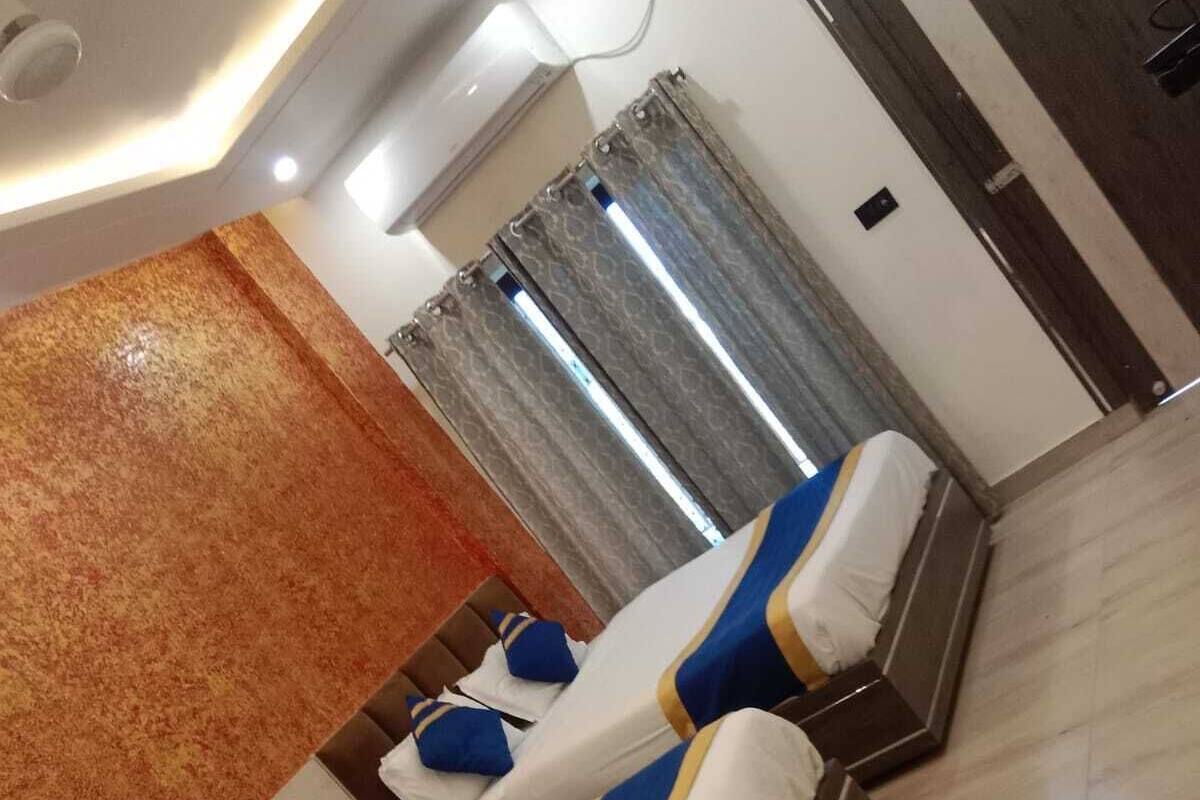 Camera d'albergo con tre letti, ognuno con biancheria bianca e blu e finiture dorate. La stanza presenta una parete di legno decorativa, un grande specchio e vista sull'esterno attraverso una finestra con tende.