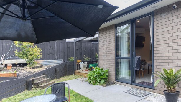 A patio area with a table and chairs, a black umbrella, and a brick building with a glass door. - New two B/room house  attached to house (Pukekohe)