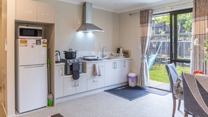 A compact kitchen with a microwave, stove, and sink. There is a small trash bin and a dining area with a table and chairs. - New two B/room house  attached to house (Pukekohe)