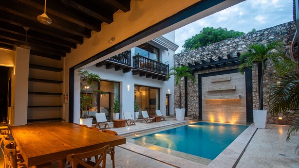Un espace aménagé au bord de la piscine, avec une table et des chaises en bois, un mur en pierre et un balcon agrémenté de plantes.