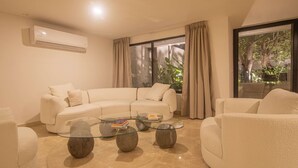 Una sala de estar moderna con un sofá seccional color beige, mesas de centro de vidrio y un ventanal con cortinas.