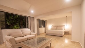 Una sala de estar moderna con un sofá color beige, una mesa de centro de vidrio y una cama con cabecera.