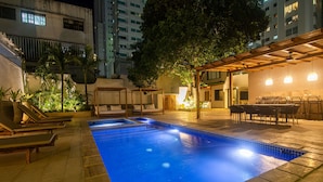 Una piscina con sillones de descanso y un área de estar cubierta, rodeada de edificios.