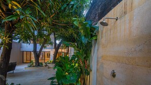 Un espacio exterior moderno con un muro de piedra, un cabezal de ducha metálico y abundante vegetación tropical.