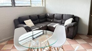 Un coin salon avec un canapé d’angle en cuir foncé, une table basse en verre et deux chaises blanches.