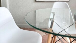 Une pièce avec une table en verre, des chaises blanches et une œuvre d’art encadrée représentant des motifs de feuilles, accrochée au mur.