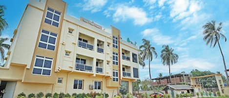 A multi-story hotel with a flat roof, balconies, and a well-maintained lawn with palm trees.