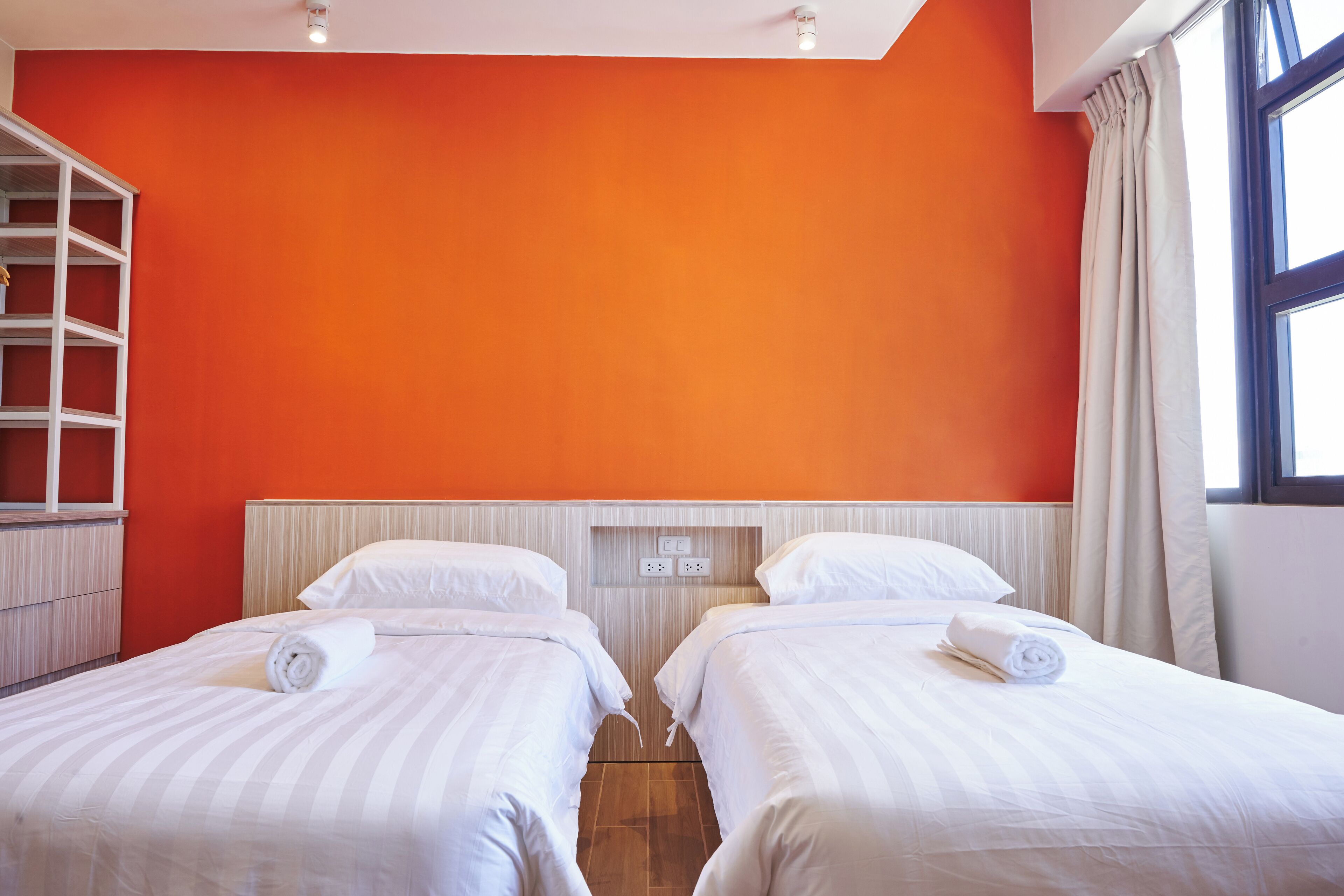A hotel room with two beds, a wooden floor, a red wall, and a window with curtains.