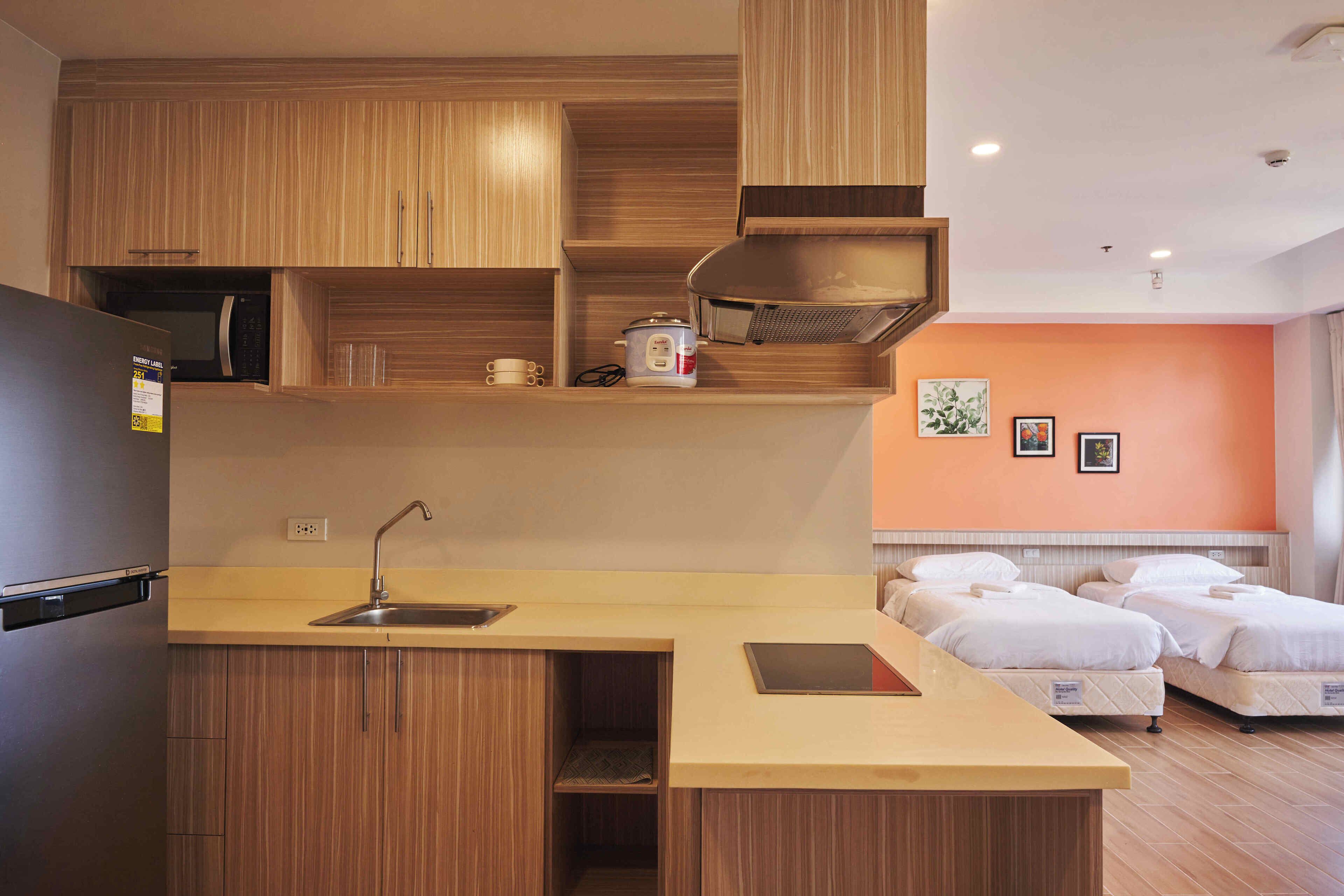 A compact kitchen with wooden cabinets, a sink, and a microwave. There are two beds with white bedding in the adjacent room.