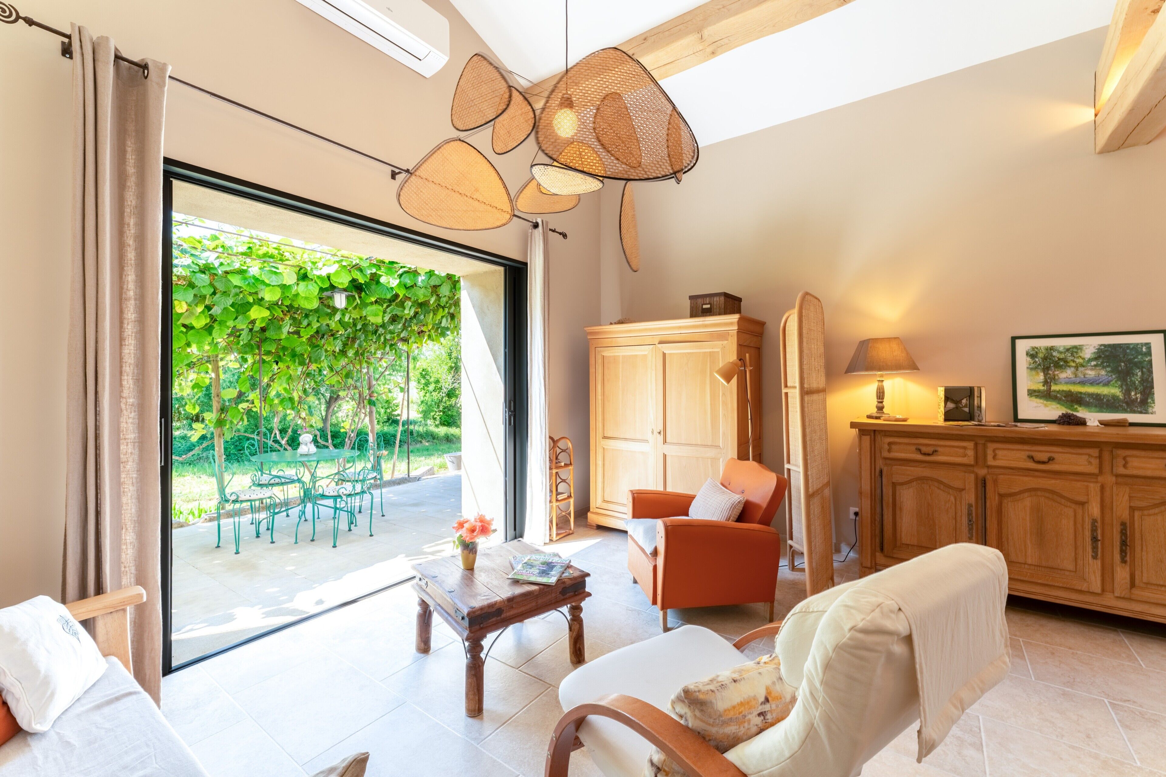 A spacious living room with a large sliding glass door leading to a garden, a wooden cabinet, a sofa, and a table with a lamp.