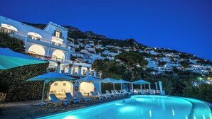 A pool area with lounge chairs and umbrellas, a building with a balcony, and a hillside with white buildings in the background.