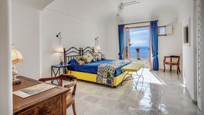 A bedroom with a bed, a desk, and a chair. There is a balcony with a view of the sea.