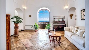 A spacious living room with a piano, a white sofa, and a view of the sea through an arched doorway.