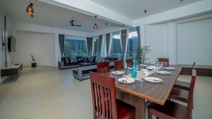 Een ruime eethoek met een lange houten tafel voor zes personen, omringd door houten stoelen. Achtergrond heeft een bank en een kleine tafel, met grote ramen die uitzicht bieden op groen buiten.