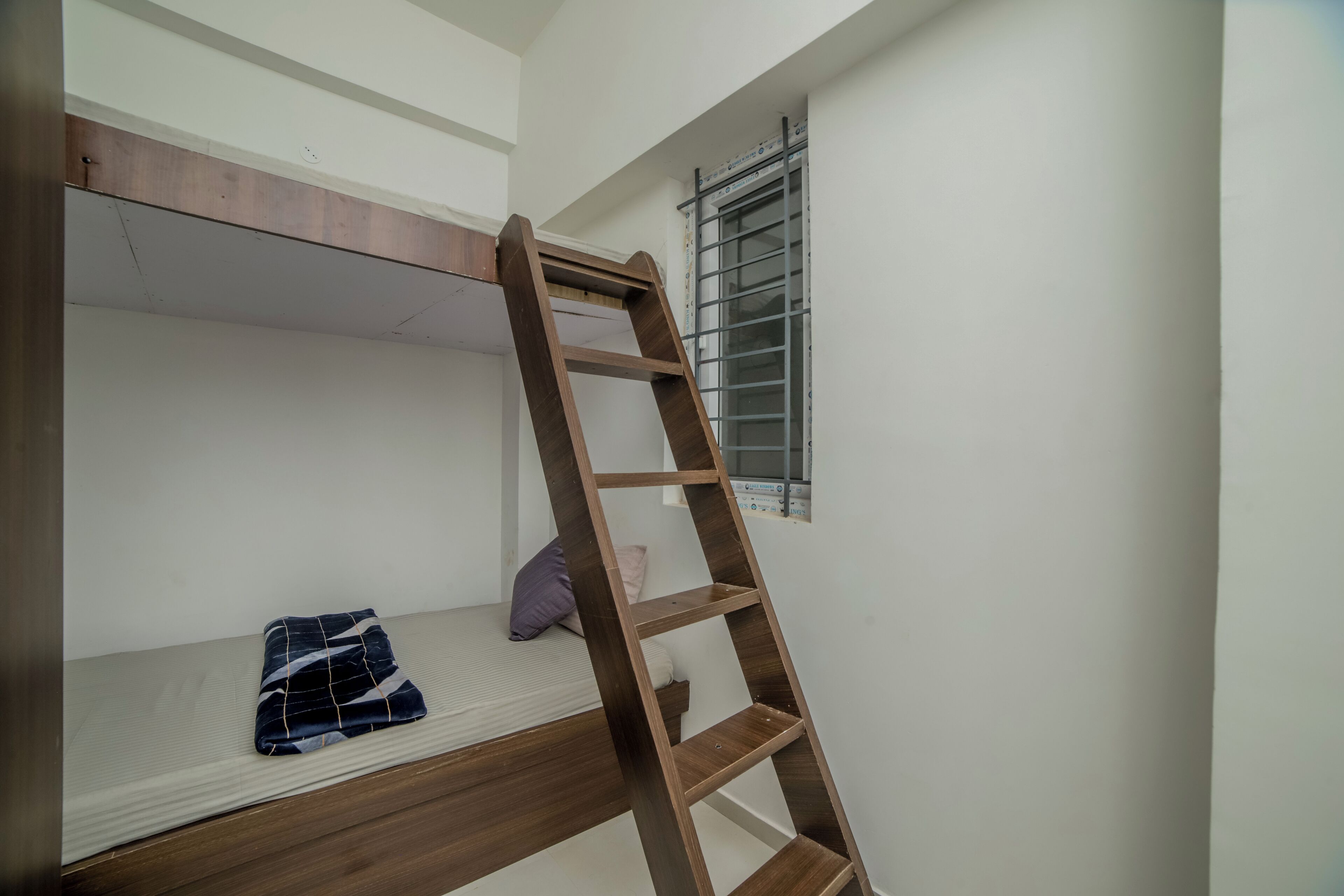 Een stapelbed met een houten trapje, een klein nachtkastje en een raam met metalen tralies.