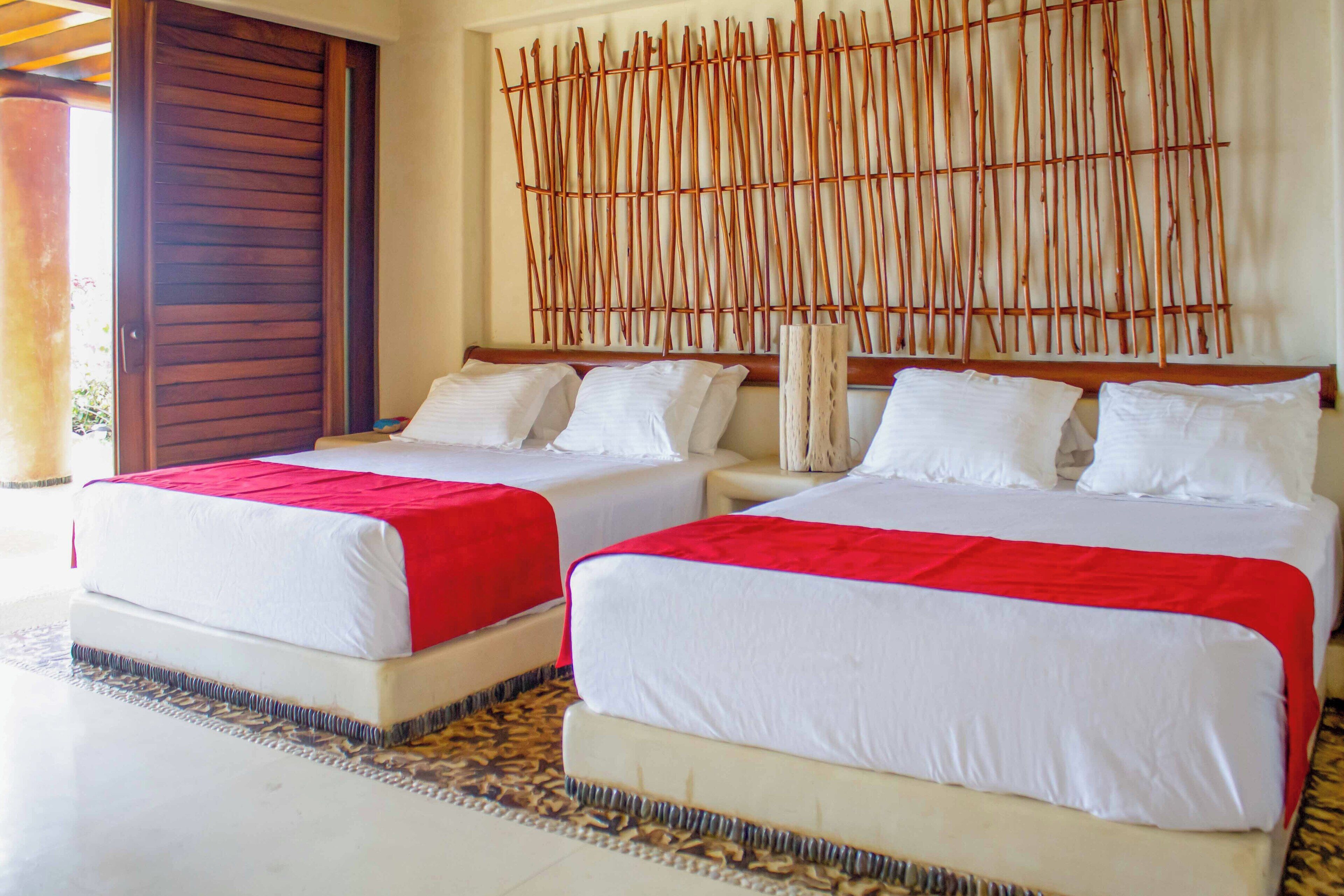 Habitación de hotel con dos camas, cada una con ropa de cama blanca y detalles rojos, un cabecero de madera y un piso con estampado.