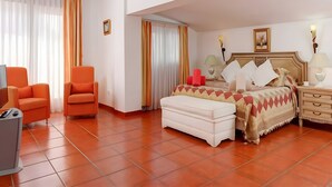 Une chambre d’hôtel avec un lit, deux fauteuils orange, un pouf blanc, une table de chevet avec une lampe et une poubelle.