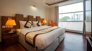 A modern hotel room with a large bed, two bedside lamps, a nightstand, and a view of the city through large windows. - HOTEL PALM D'OR (New Delhi)