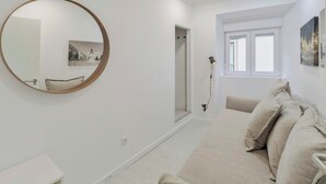 A modern, minimalist bedroom with a large round mirror, a bed with beige bedding, a bedside table, and a window with natural light.