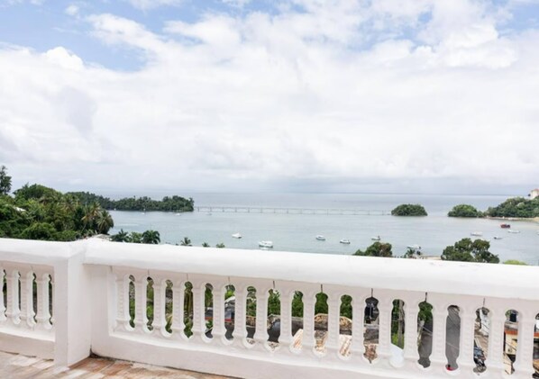 Un balcon doté d’une balustrade blanche, offrant une vue sur la côte, avec des bateaux et un pont.