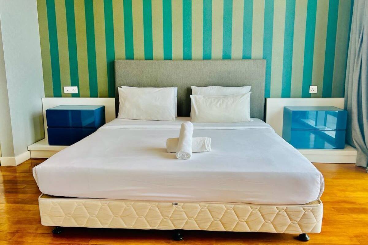 A neatly made bed with white linens and a quilted mattress, two pillows, and a folded towel in the center. There are two bedside tables with blue storage boxes, and the room has a striped wall and wooden flooring.