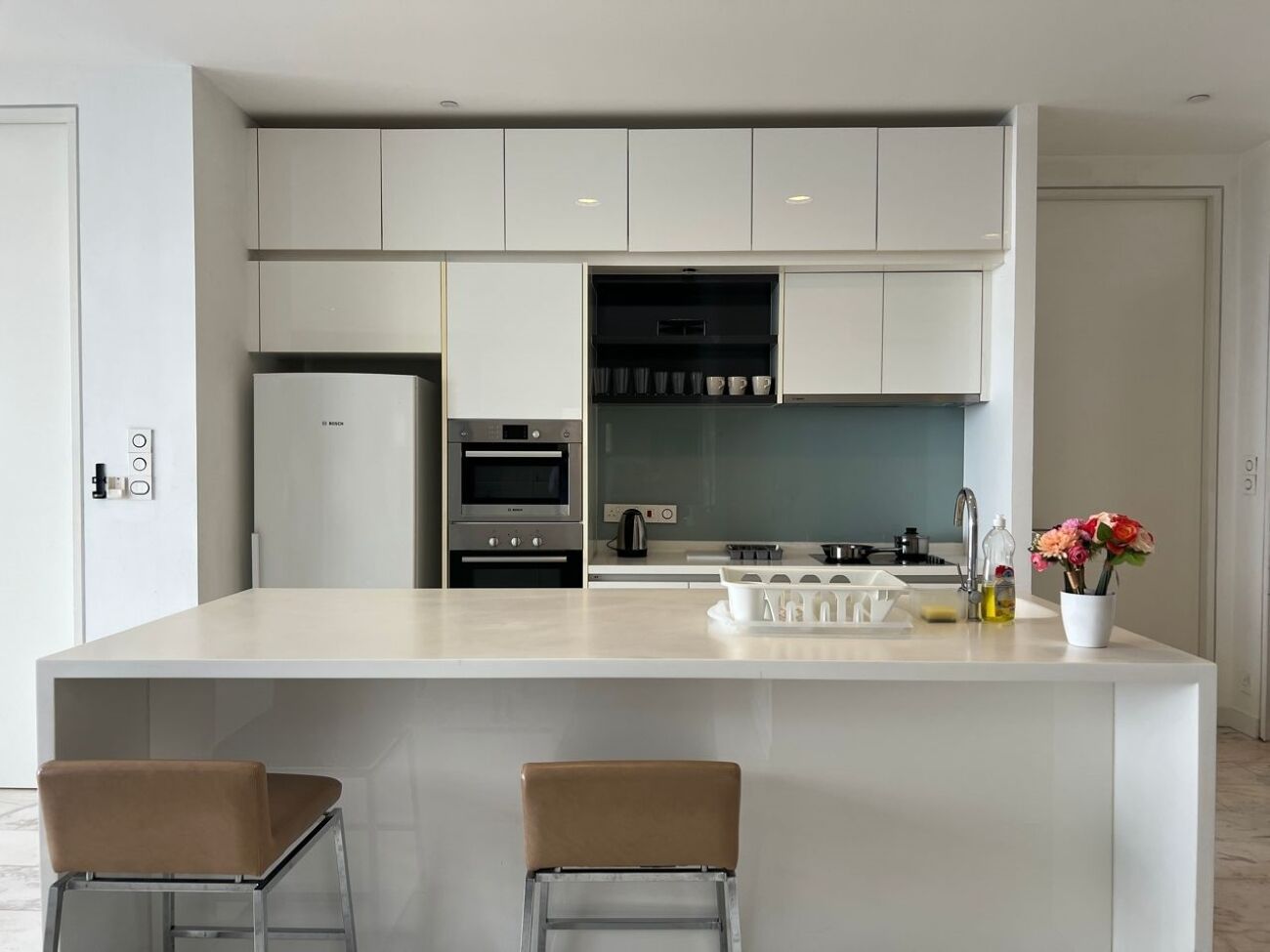 Cocina moderna con armarios blancos, una isla central con taburetes de bar, electrodomésticos integrados y una pequeña zona de encimera con un jarrón de flores.