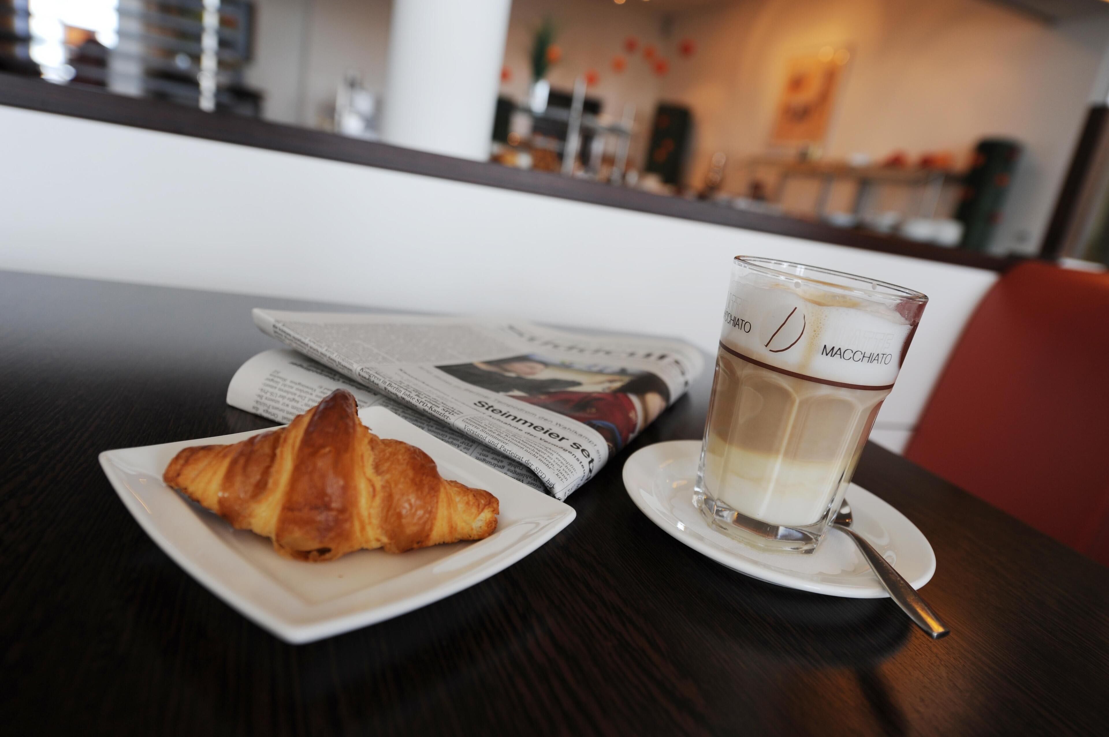 A croissant on a white plate, a glass of coffee, and a newspaper on a table in a cafe setting.
