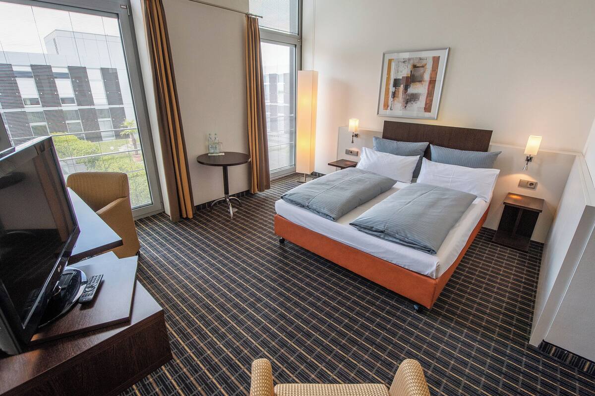 Ein Hotelzimmer mit einem großen Bett, einem Schreibtisch mit Fernseher und Fernbedienung, einem kleinen Tisch und Blick auf Gebäude.
