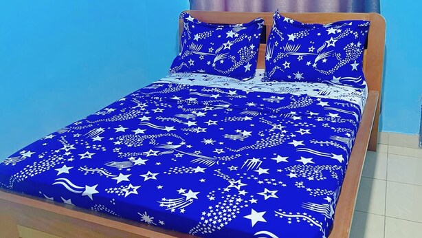 Una cama con una colcha azul estampada y almohadas a juego, un cabecero de madera y una cortina.
