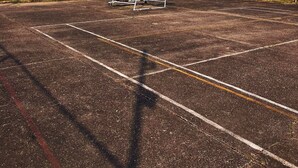 Una cancha de baloncesto al aire libre con un aro dañado, rodeada de edificios y palmeras.