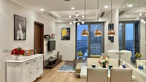 A modern living room with a flat-screen TV, a dining area with a table and chairs, and a large window with a city view. - HMH- Vinhomes TIme City - Park Hill  (Hanoi)