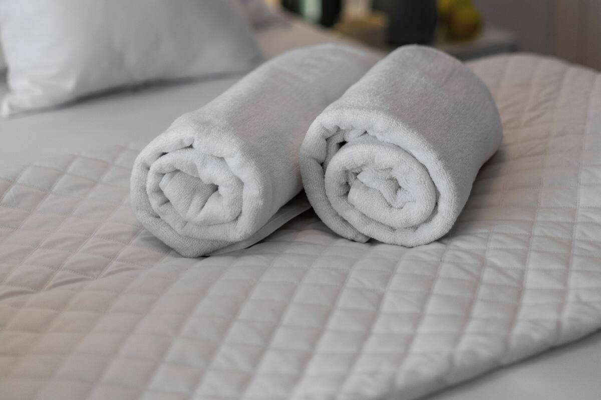 A neatly made bed with rolled white towels, a white pillow, and a bedside lamp with a vase of flowers.