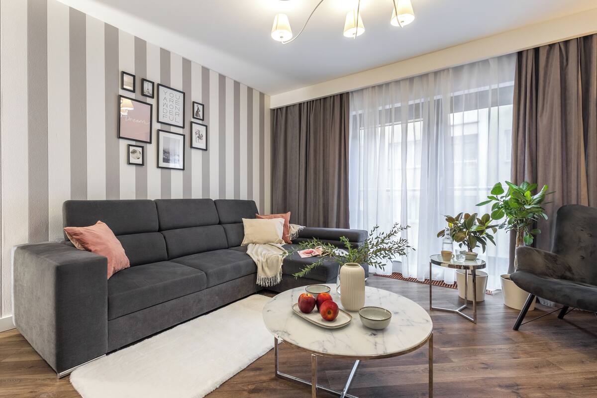 A modern living room with a dark grey sofa, a round coffee table with apples and a vase, and a striped wall with framed pictures.