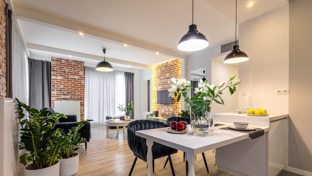 A modern kitchen with white countertops, a dining area with a black chair, and a brick wall. - Absyntapart Krawiecka - parking w cenie (Wroclaw)