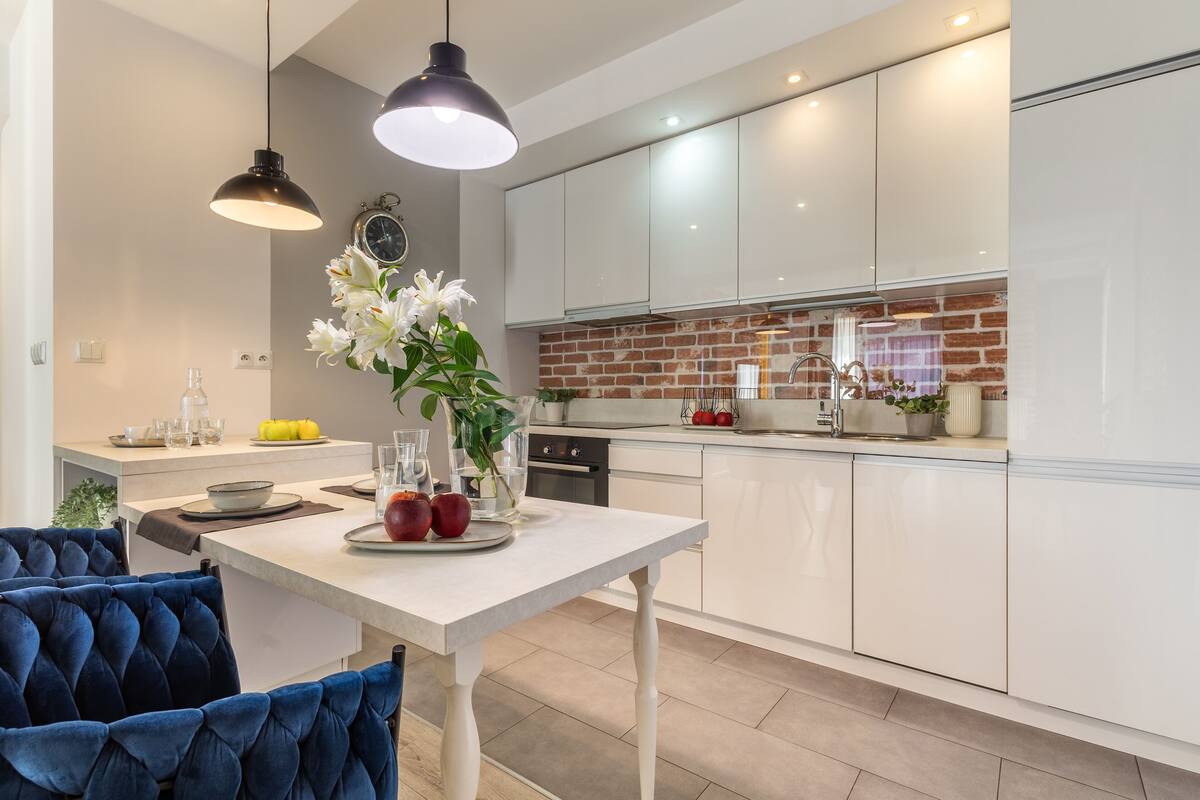 A modern kitchen with white cabinets, a brick backsplash, and a dining area with a white table and blue upholstered chairs.