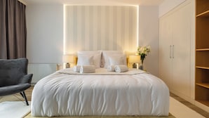 A neatly made bed with white linens, a grey armchair, and a bedside table with a lamp and a vase of flowers. - Absyntapart Krawiecka - parking w cenie (Wroclaw)