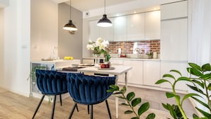 A modern kitchen with white cabinets, a brick backsplash, and a dining area with two blue velvet chairs and a white table. - Absyntapart Krawiecka - parking w cenie (Wroclaw)