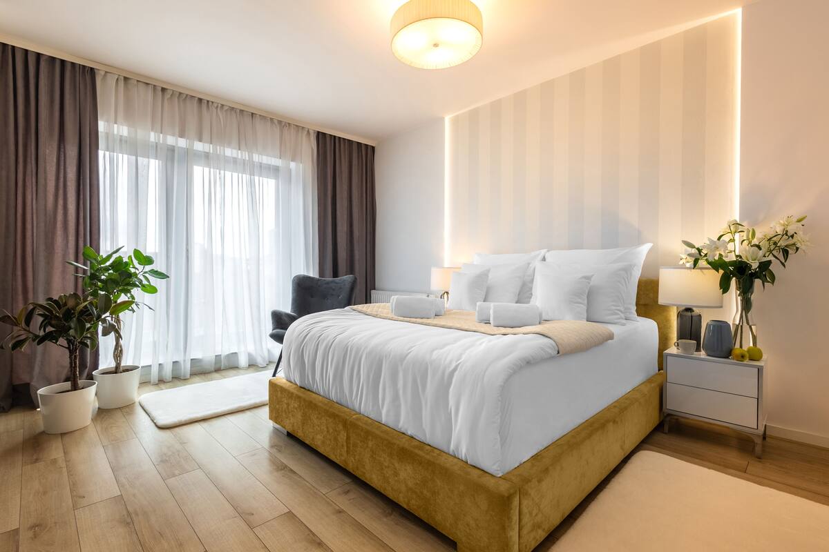 A modern hotel room with a large bed, bedside tables, a nightstand, and potted plants.