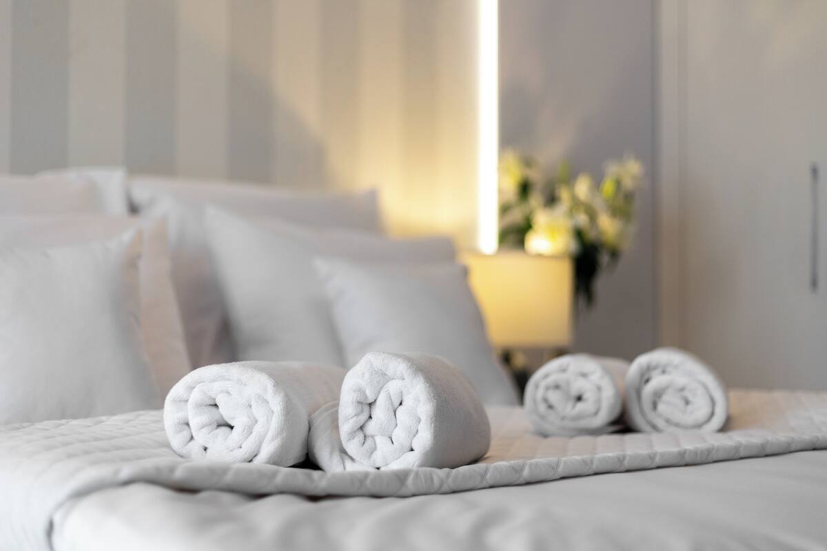 A neatly made bed with rolled white towels, a white quilted blanket, and a bedside lamp with a floral arrangement.