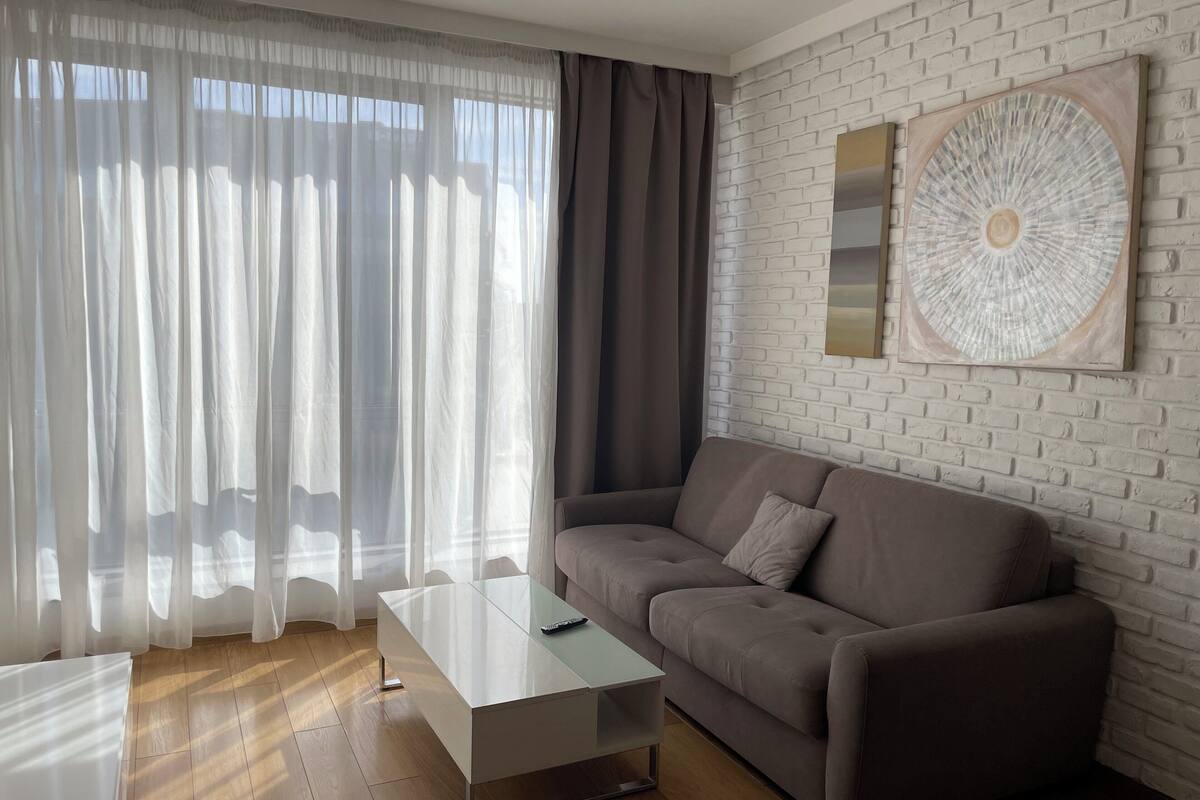 A modern living room with a sofa, a glass coffee table, and a large window with sheer curtains.