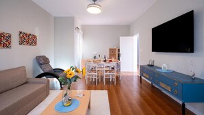 Een woonkamer met een bank, een eettafel voor vier personen, een aan de muur bevestigde televisie en een blauwe kast.