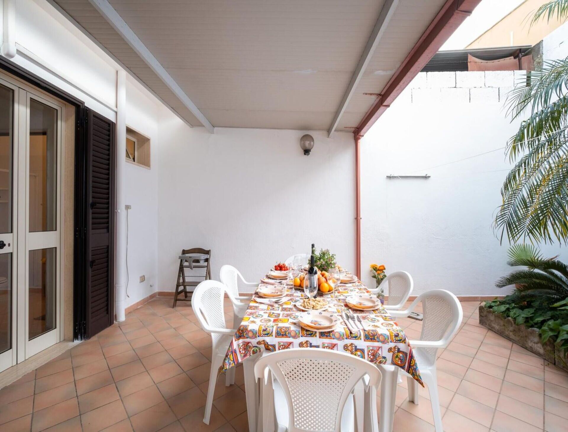 Un patio techado con una mesa preparada para una comida, rodeado de sillas blancas y un piso de baldosas.