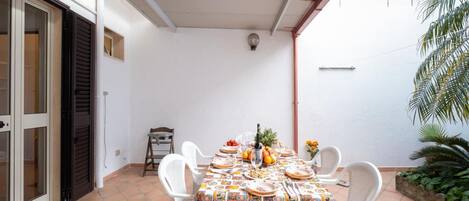 Un patio techado con una mesa preparada para una comida, rodeado de sillas blancas y un piso de baldosas.