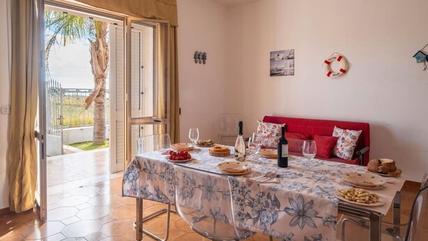 A dining area with a table set for two, a red sofa, and a view of a palm tree through an open door.